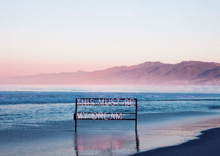 a wooden sign sitting on top of a beach next to the ocean with mountains in the background