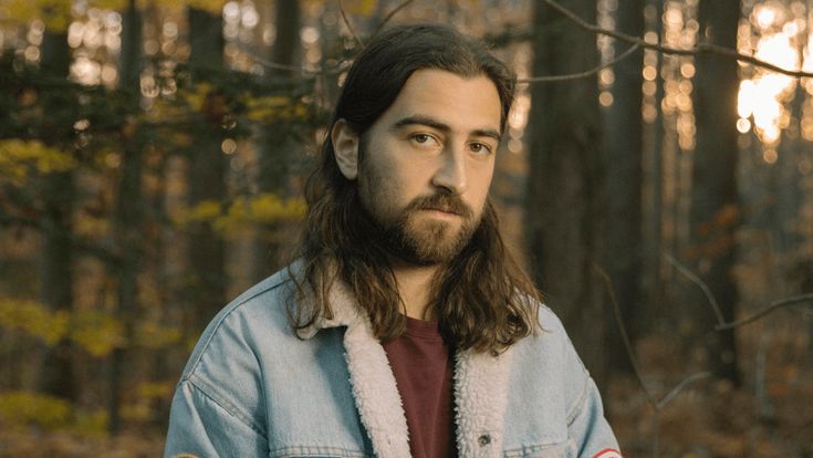 a man with long hair and beard wearing a jean jacket standing in front of trees