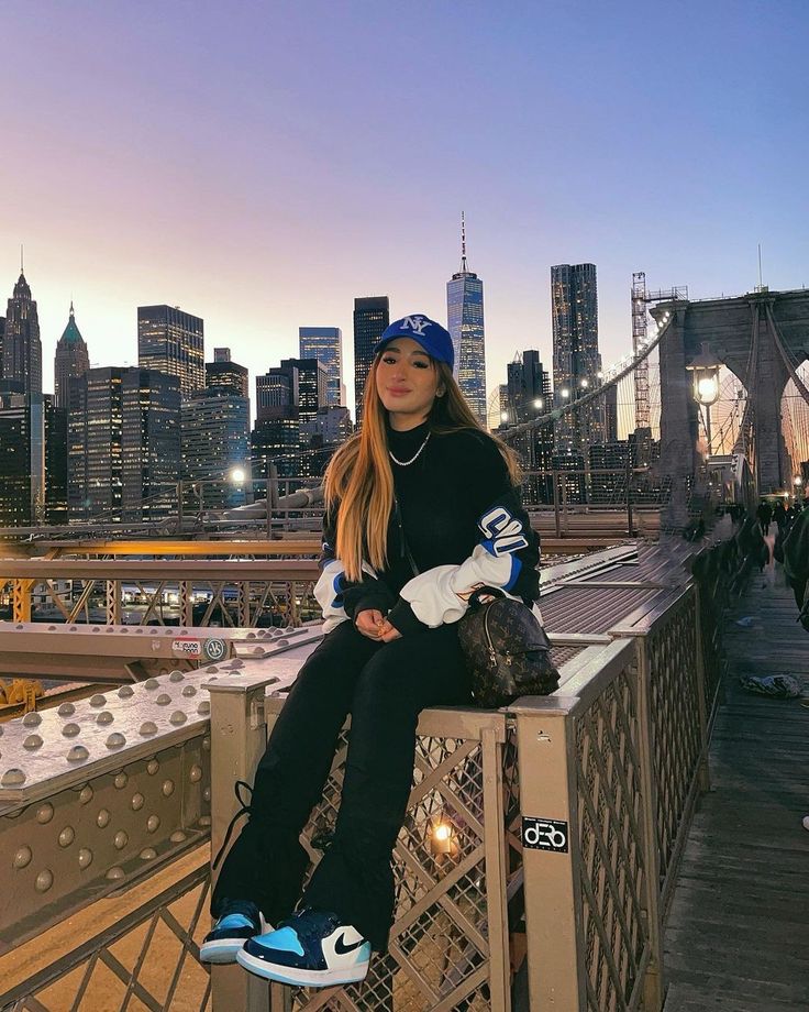 a woman sitting on top of a metal fence next to a bridge with the city skyline in the background