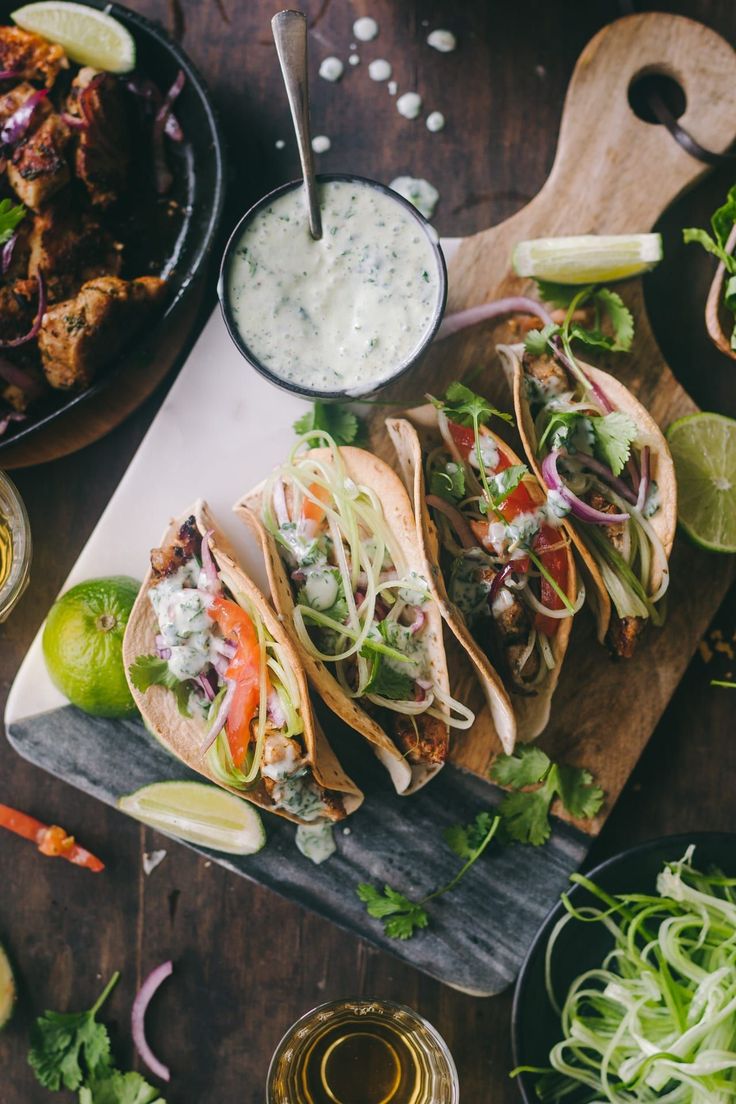 several tacos are lined up on a cutting board with limes, cilantro, and sour cream