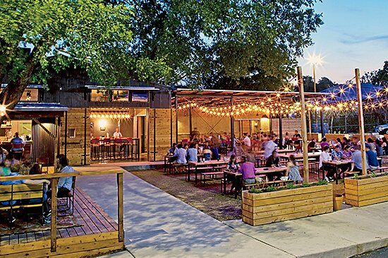 people are sitting at tables and eating outside in the evening time with lights strung over them