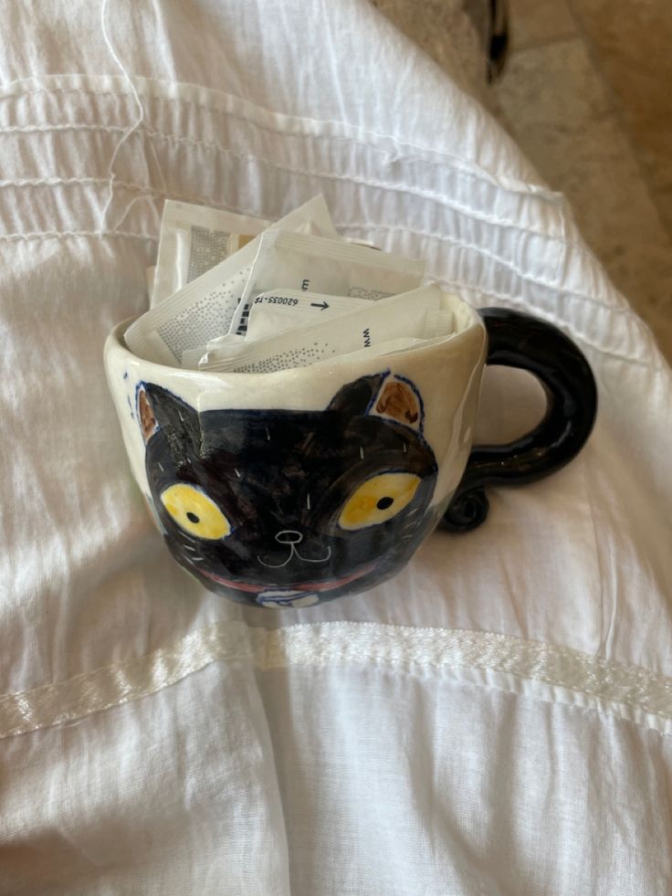 a black cat mug sitting on top of a white bed covered in sheets and pillows