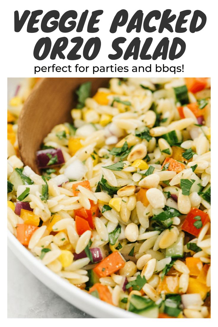 vegetable packed orzo salad in a white bowl with a wooden spoon on the side