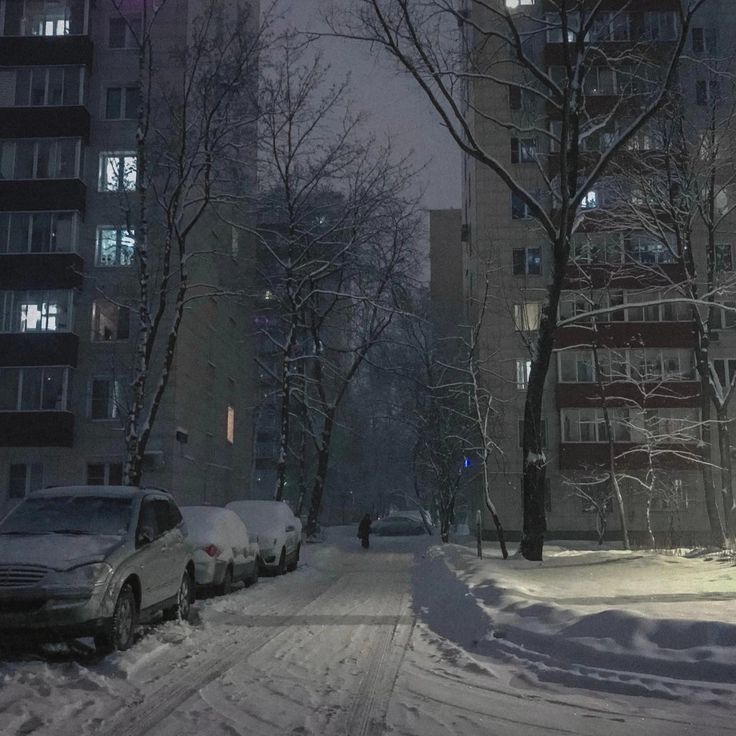 cars parked on the side of a snow covered road in front of tall buildings at night