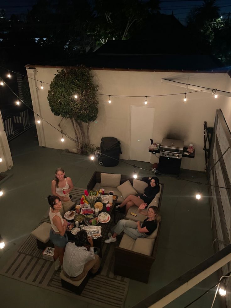 a group of people sitting around a table on top of a patio with lights strung from the ceiling