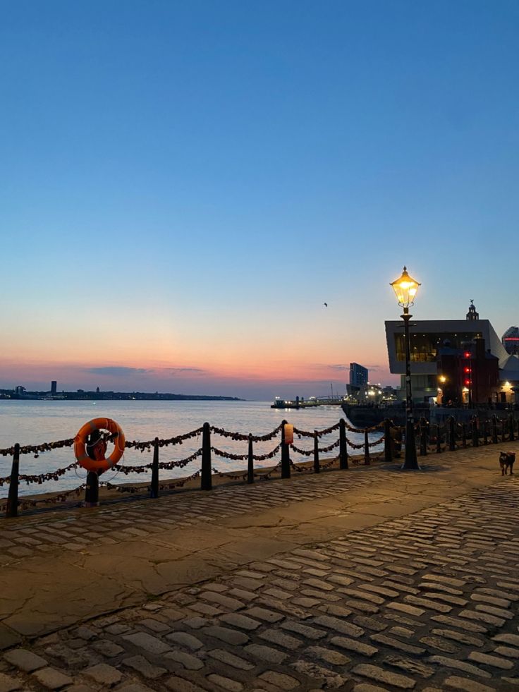 a brick walkway next to the water at dusk with lights on and a boat in the distance