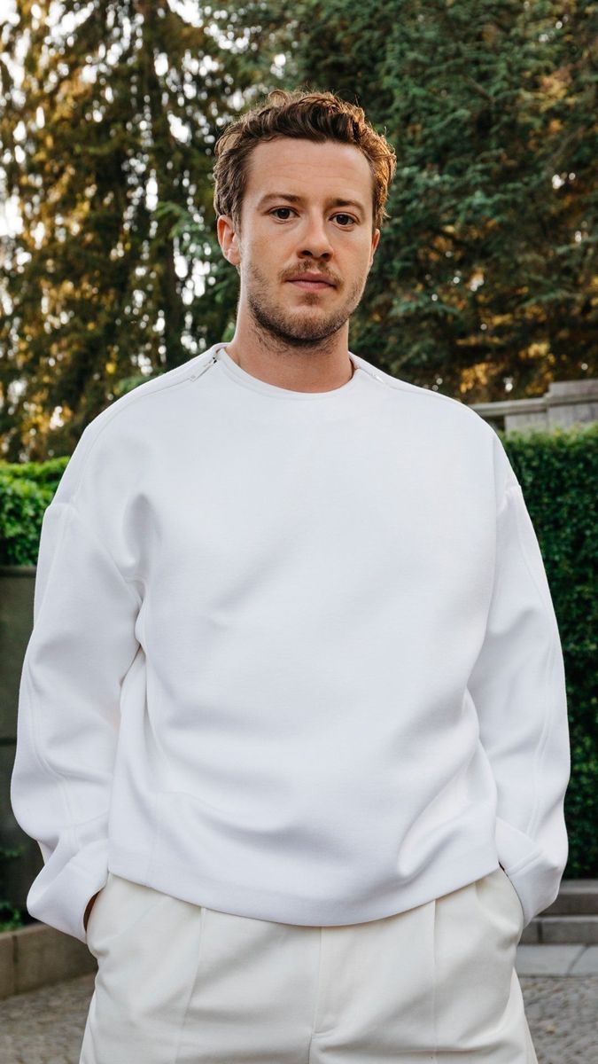 a man standing in front of some trees wearing a white sweatshirt and pants with his hands in his pockets