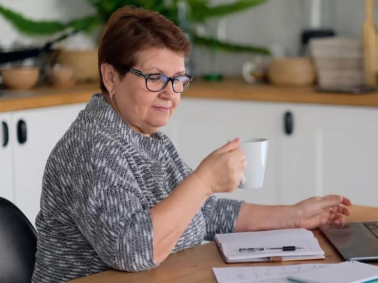 a woman sitting at a table with a laptop and coffee mug in her hand, looking off to the side