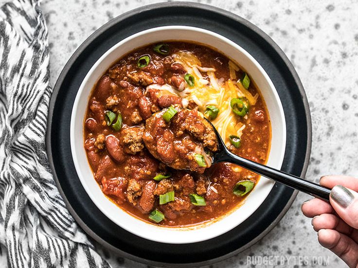 a person holding a spoon in a bowl of chili and meat stew with cheese on top