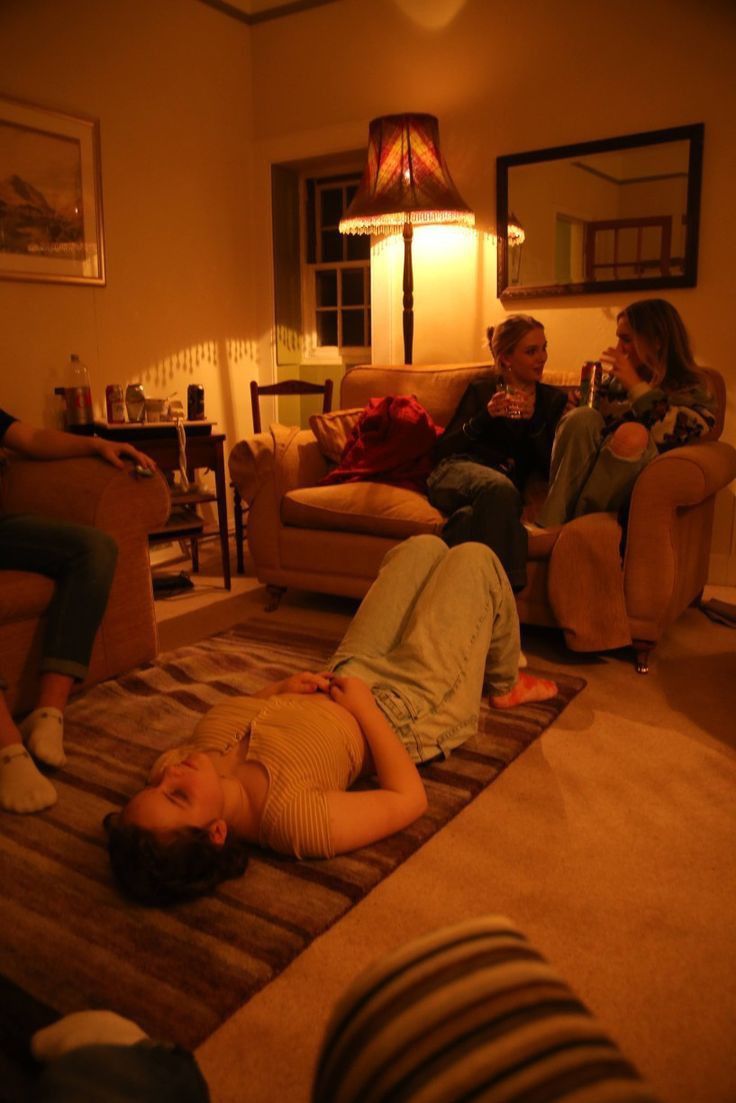 three people sitting in a living room with one laying on the floor and another standing