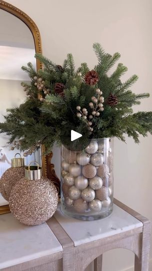 a vase filled with christmas balls and greenery on top of a table next to a mirror