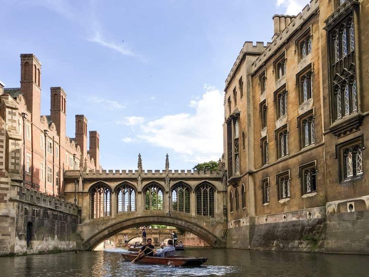 two people in a small boat on the water near some buildings and a bridge with arched windows