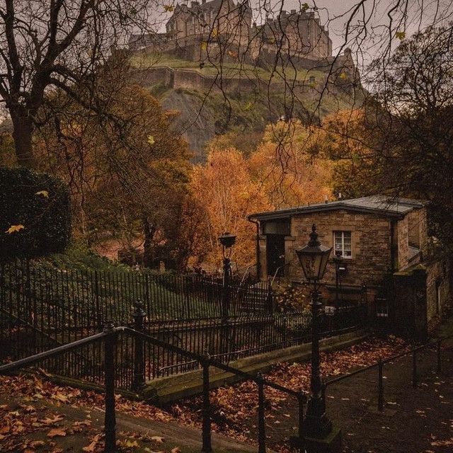 an old stone house in front of a castle with autumn leaves on the ground and trees around it