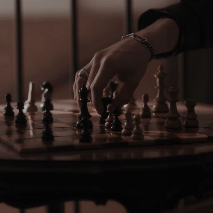 a person is playing chess on a table with other pieces in the foreground and one hand reaching for it