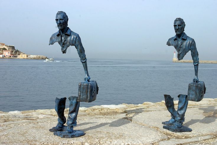 two statues of men carrying bags on their backs near the water with buildings in the background