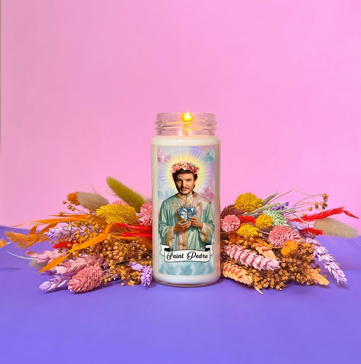 a candle with the image of jesus on it surrounded by flowers and leaves against a pink background