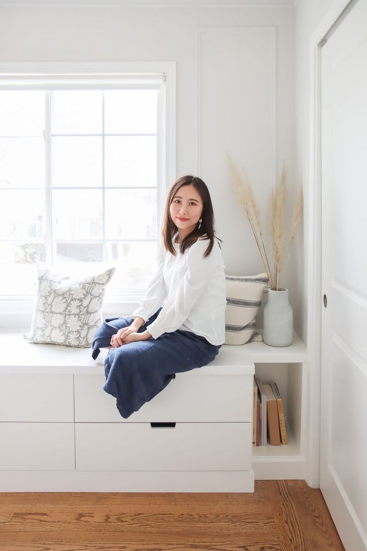 a woman sitting on top of a window sill next to a book shelf and pillows