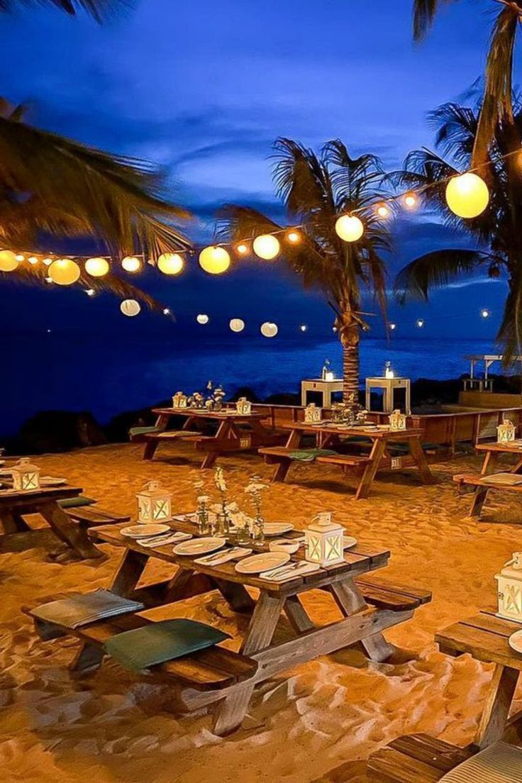 an outdoor dining area on the beach with lights strung from palm trees and hanging lanterns