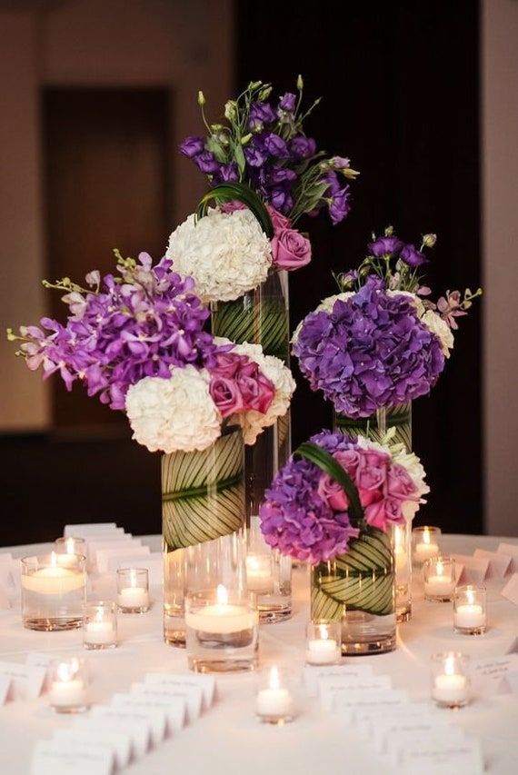 purple and white flowers are in vases on a table with votive lit candles