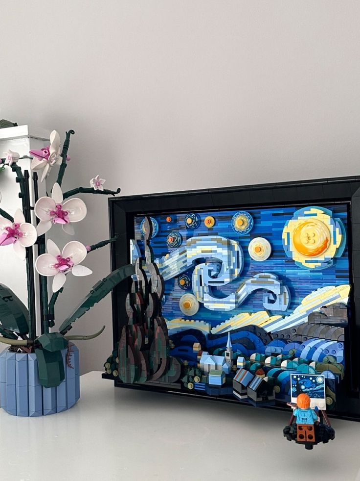 a vase with flowers next to a painting on the wall and a lego figure sitting in front of it