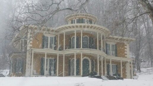 a large house in the middle of winter with snow falling on it and trees around