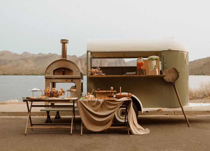 an old camper is parked next to a lake with food on the table in front of it