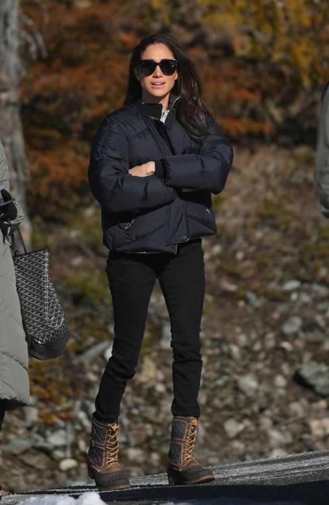 a woman in black jacket and sunglasses standing on snow covered ground with her arms crossed