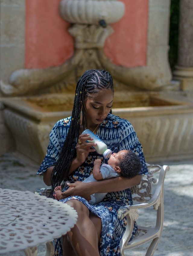 a woman holding a baby while sitting on a bench in front of a water fountain