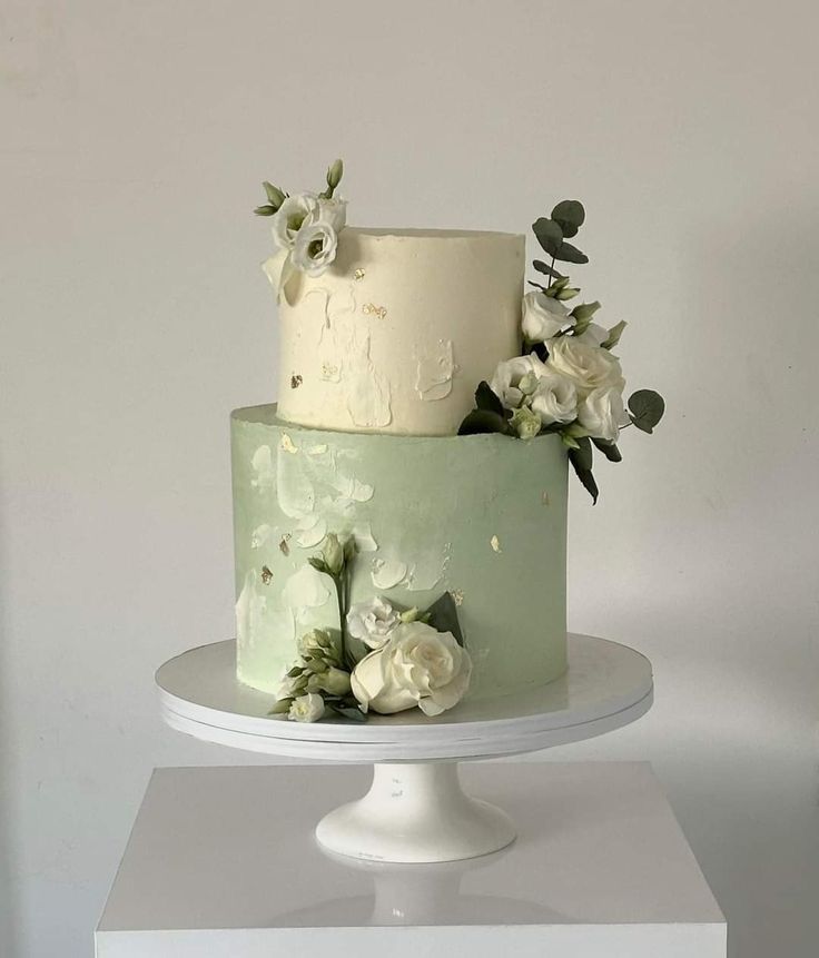 a three tiered cake with white flowers and greenery on the top is sitting on a pedestal