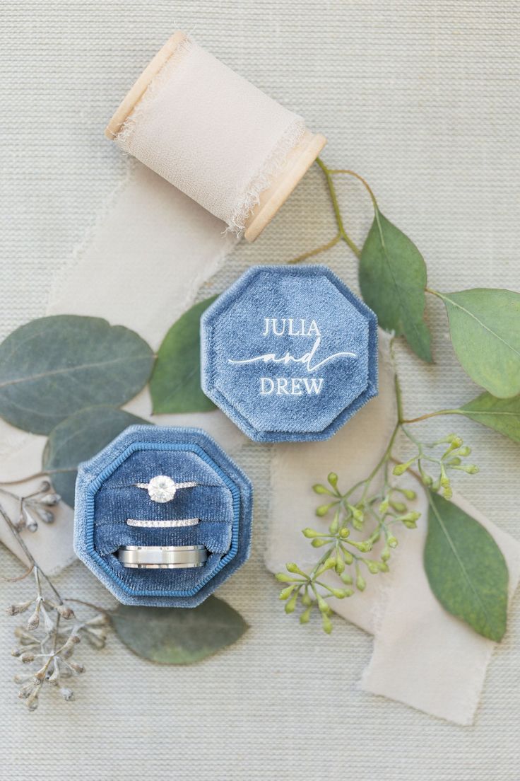 two wedding rings sitting in an open ring box next to greenery and napkins