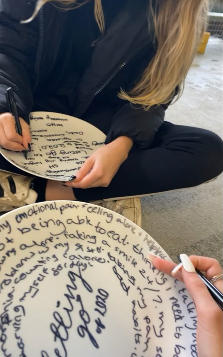 a woman sitting on the ground with two plates in front of her and writing on them