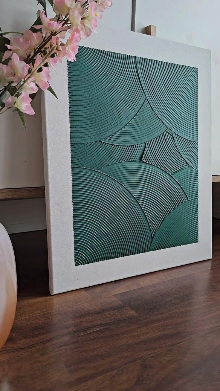 a vase filled with pink flowers sitting next to a white framed art piece on top of a wooden floor