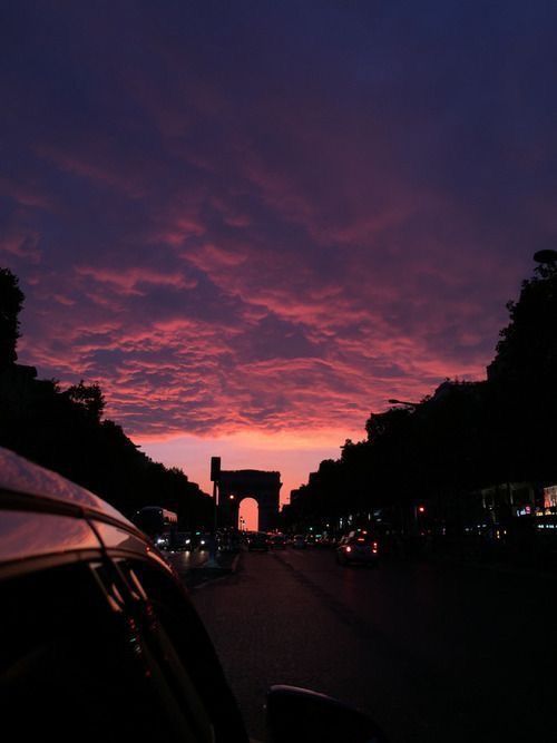 the sun is setting over an arch in the distance, with cars parked on the side of the road