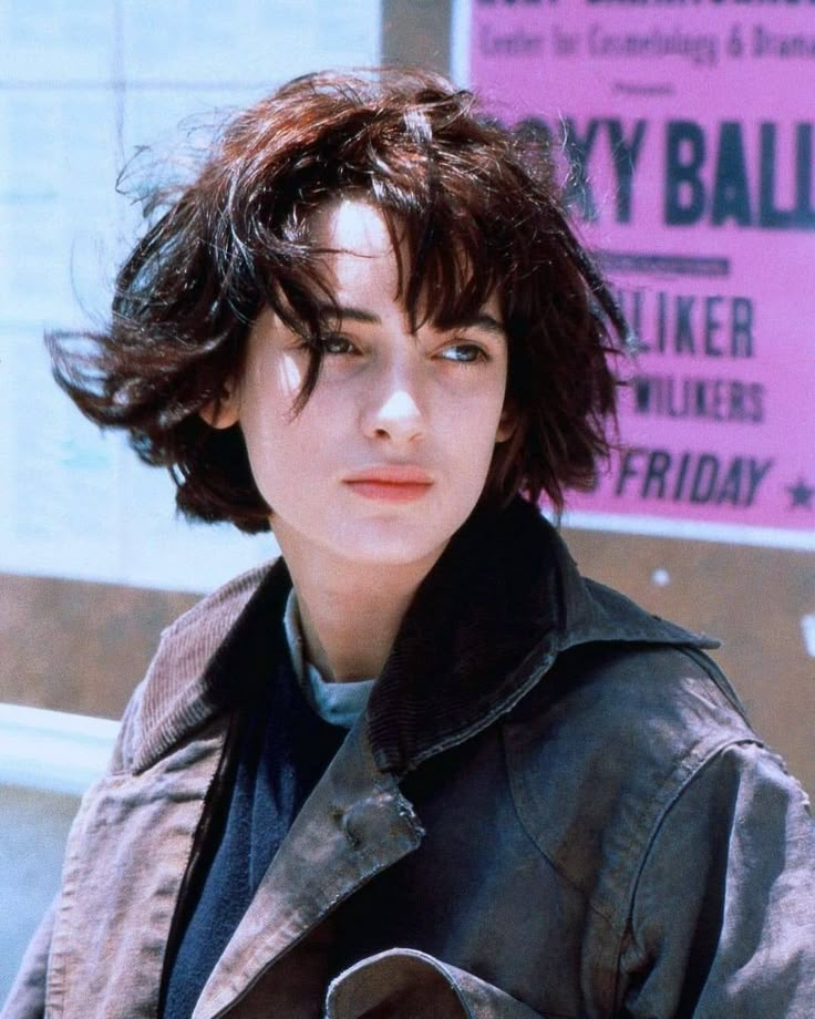 a woman with short hair standing in front of a pink sign and wearing a leather jacket