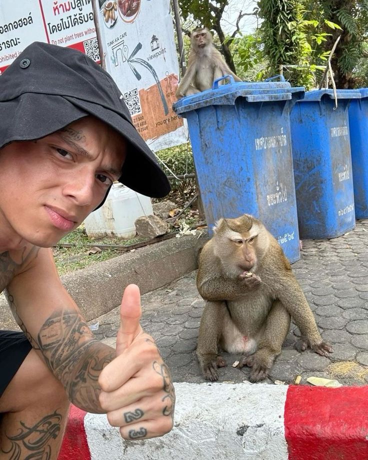 a man giving the thumbs up while sitting next to a monkey in front of some trash cans