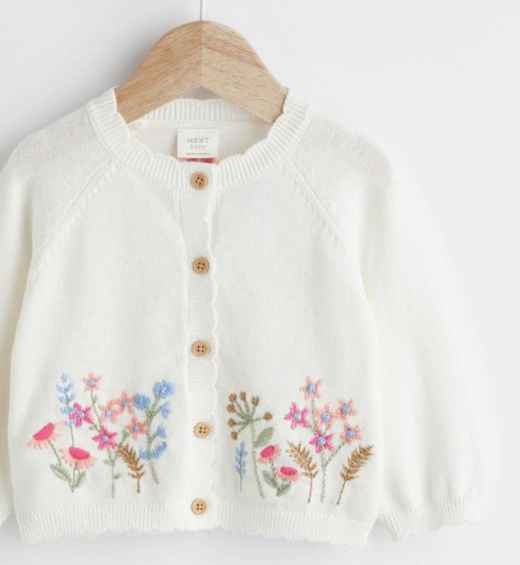 a white cardigan sweater with flowers on the front and buttons at the back, hanging from a wooden hanger