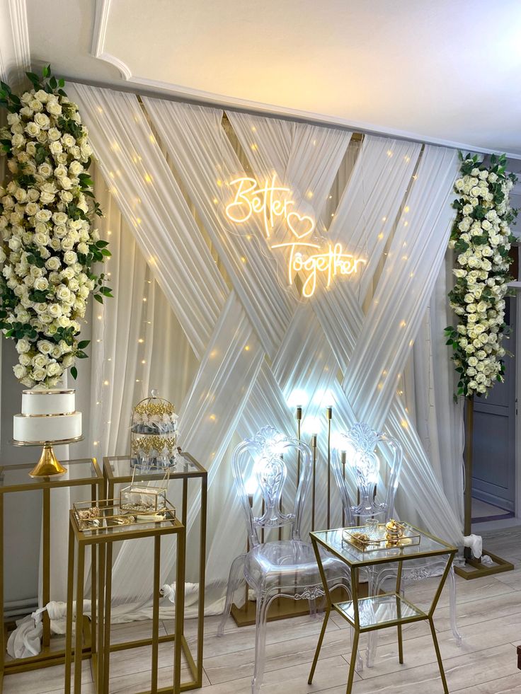 a white and gold wedding set up with flowers on the wall, chairs and table