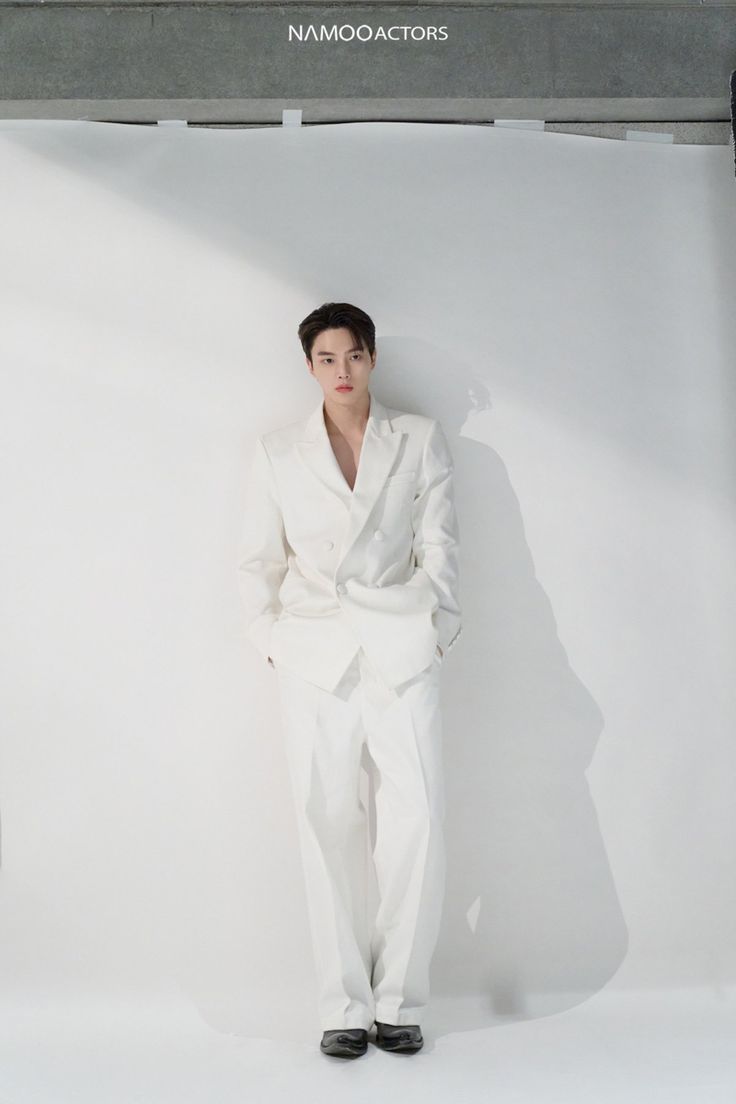 a male in a white suit and black shoes standing next to a white wall with the name namboccators on it