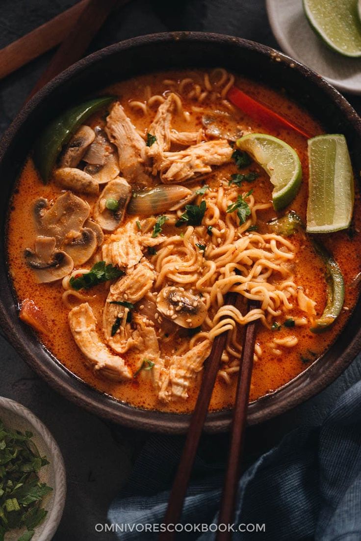 an overhead view of a bowl of noodles with chicken and mushrooms, garnished with sliced limes