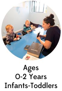 a woman sitting at a blue table with two small children in front of her and an adult holding a cell phone