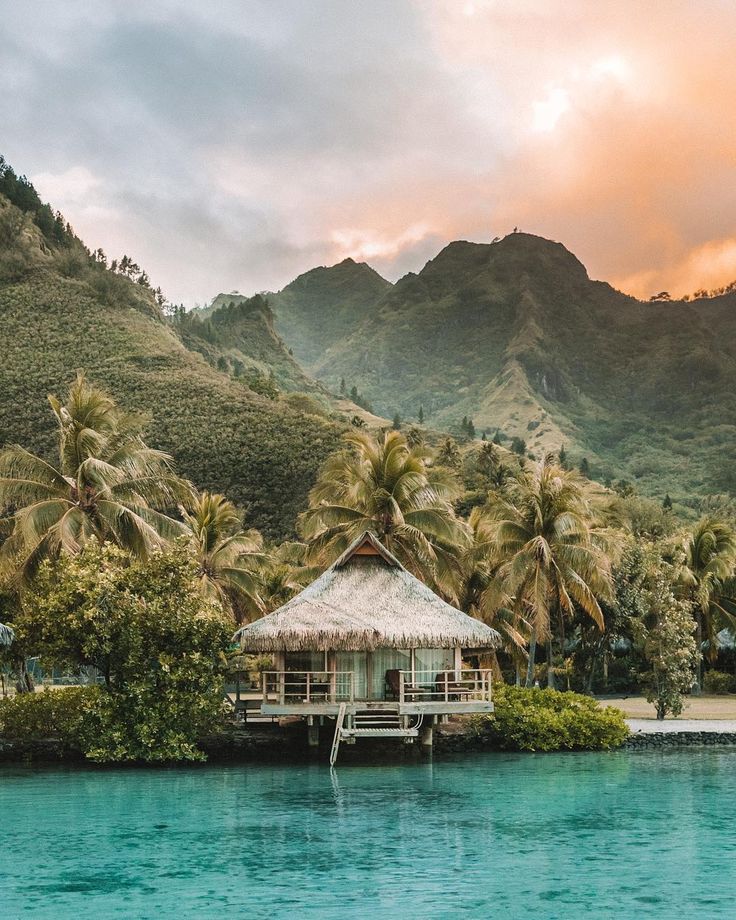 a hut on an island in the middle of water with palm trees and mountains in the background