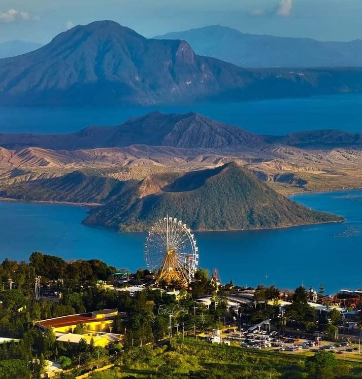 a ferris wheel on top of a hill next to water and mountains in the background