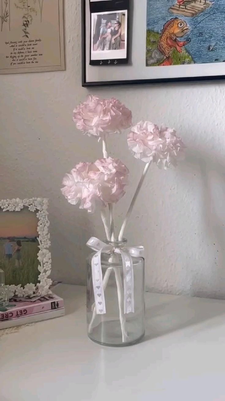 pink carnations in a clear glass vase on a white table with framed pictures