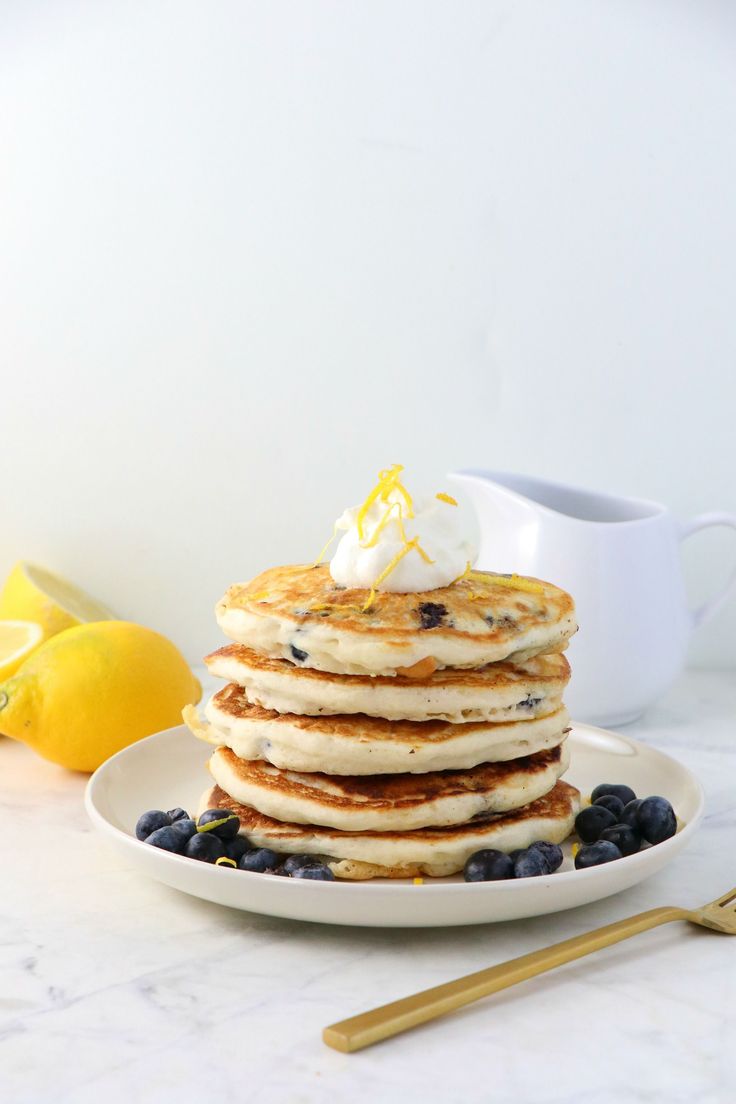 a stack of blueberry pancakes on a white plate with lemons and whipped cream