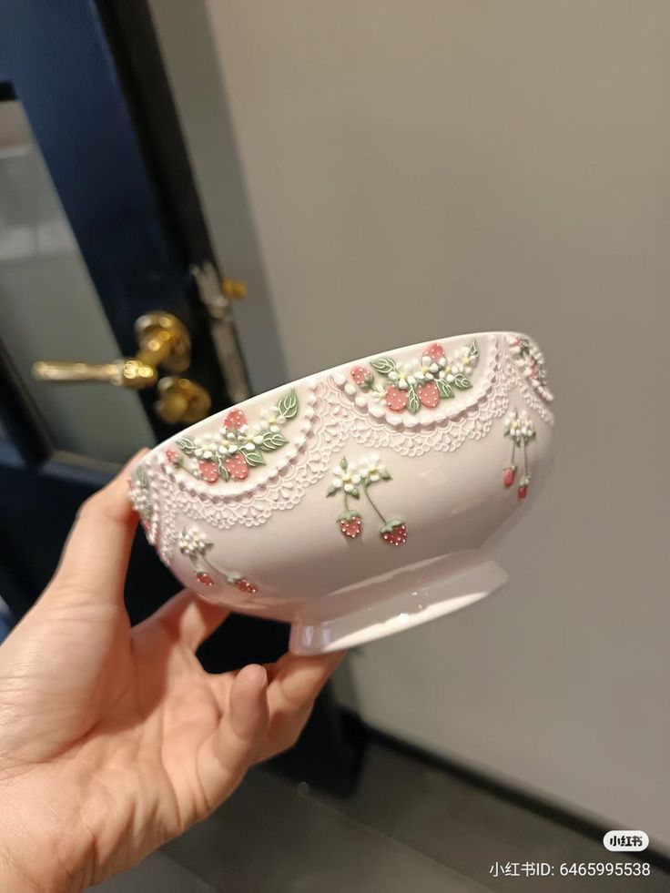 a hand holding a white vase with pink flowers on it and lace trimming around the edges