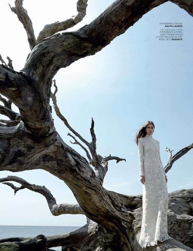 a woman in a white dress standing next to some dead trees by the ocean with blue sky