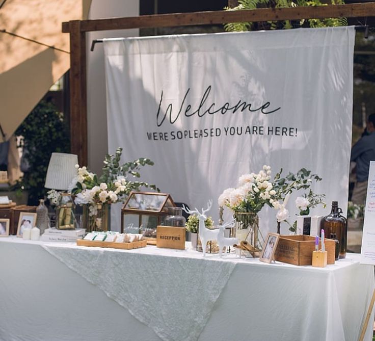 a table with flowers and personal items on it at an outdoor wedding reception in front of a welcome sign