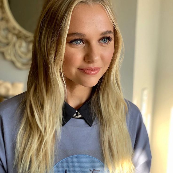 a woman with long blonde hair wearing a blue shirt and looking at the camera while standing in front of a mirror