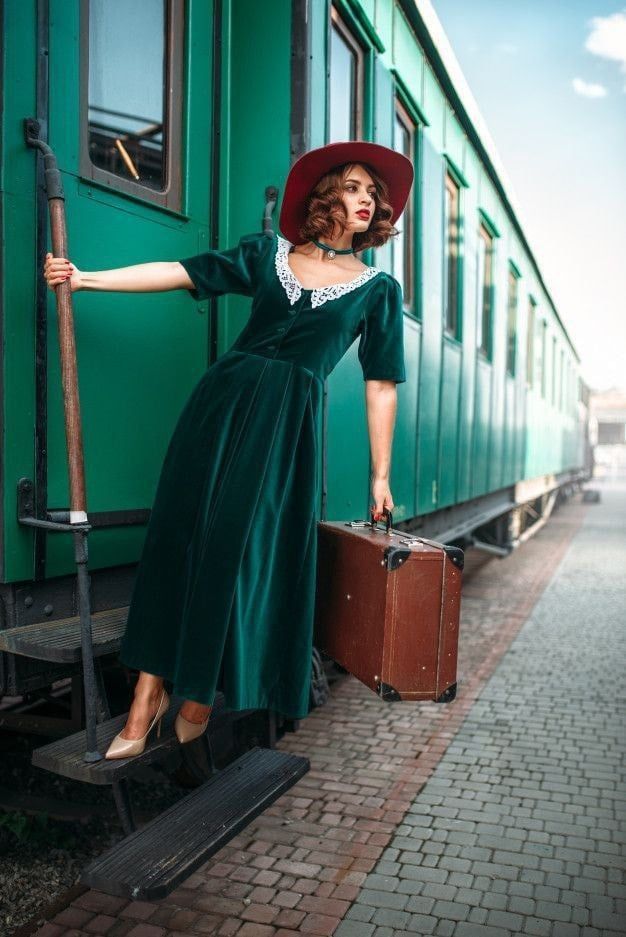 a woman in a green dress and hat leaning on a train car with her suitcase