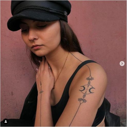 a woman wearing a black hat with tattoos on her arm and shoulder is leaning against a wall